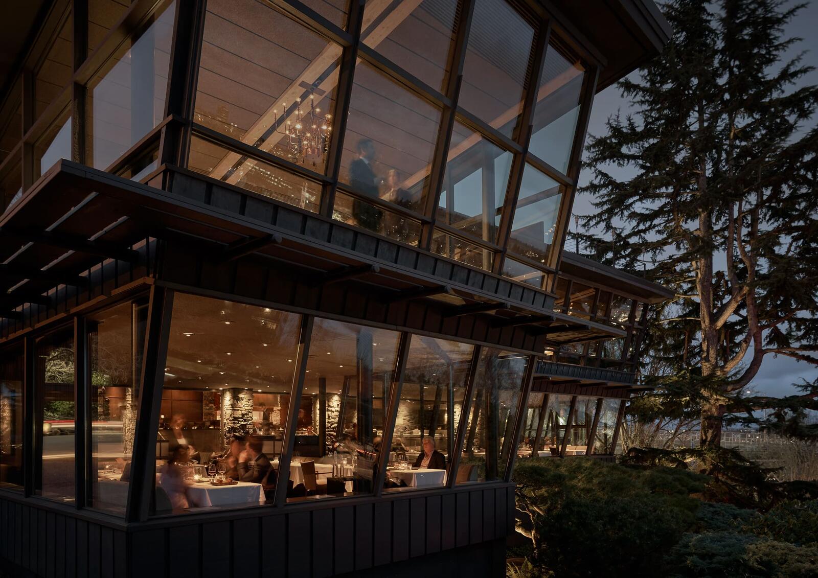 Exterior view of Canlis at night showing both floors and a bustling dining room next to a towering Lebanese Cedar